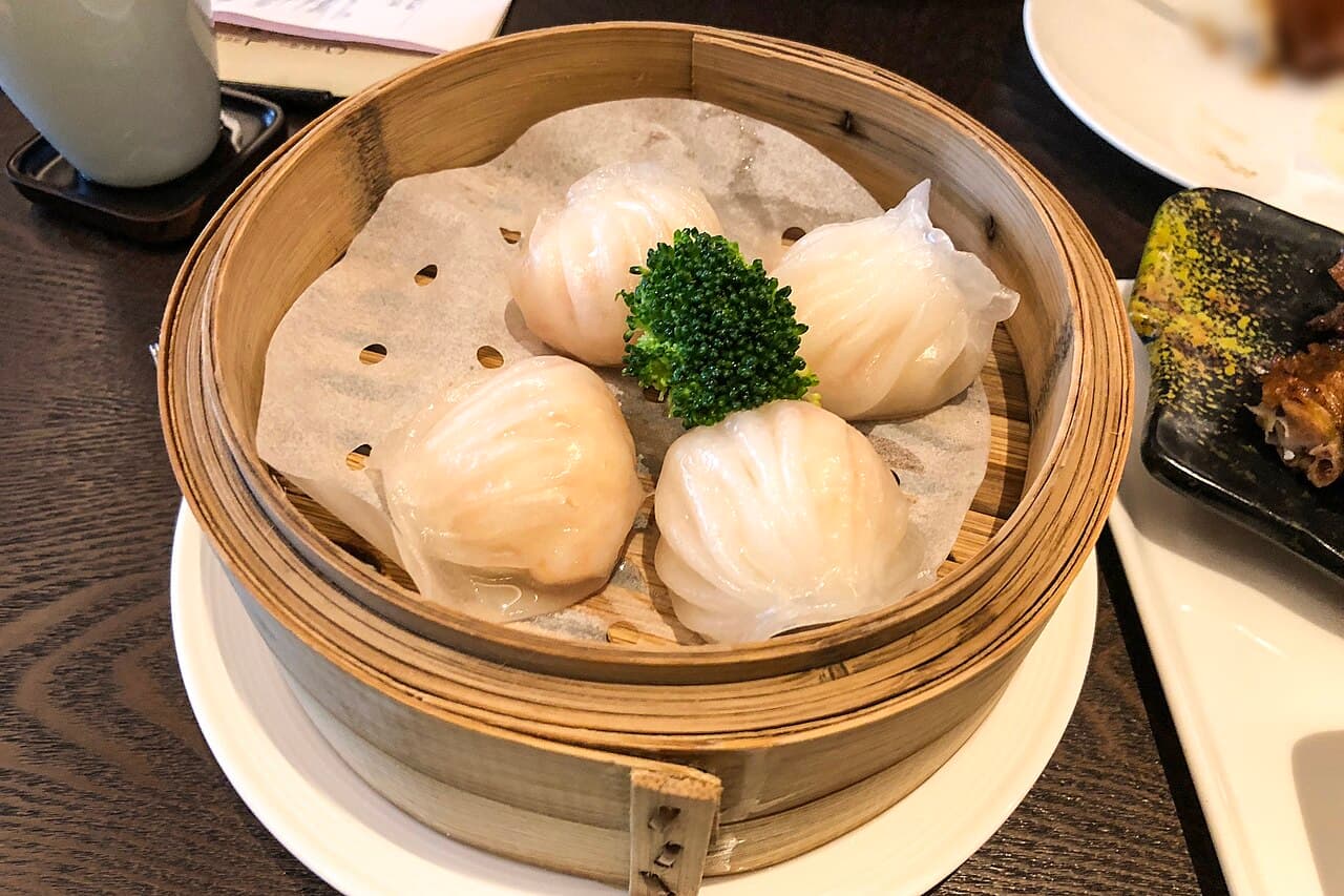 Four translucent pleated dumplings in a bamboo steamer, with pink shrimp visible through the wrappers